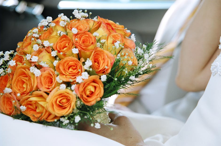 A person holding a bouquet of fresh orange roses arranged with small white filler flowers and greenery, wrapped in a transparent cellophane with a decorative golden ribbon. The bouquet is displayed ag