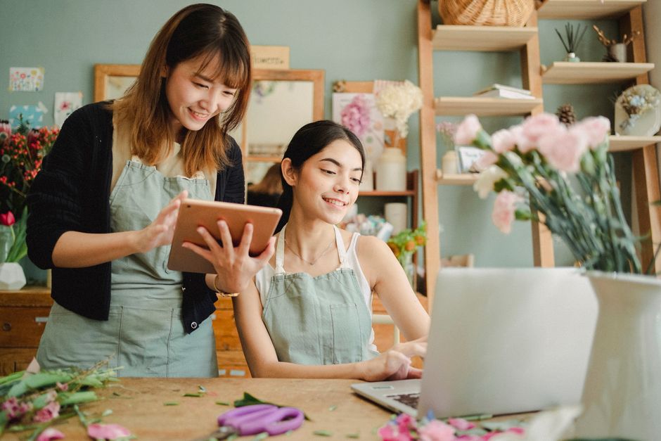Two women in a flower shop are engaged in floral arrangement work; one is standing and holding a tablet device, smiling, while the other is seated at a wooden workbench, also smiling and working on a 