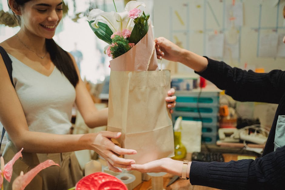 A woman with long dark hair, dressed in a white sleeveless top, is smiling as she receives a bouquet of fresh flowers from a florist. The bouquet includes elegant white calla lilies, pink roses, and g