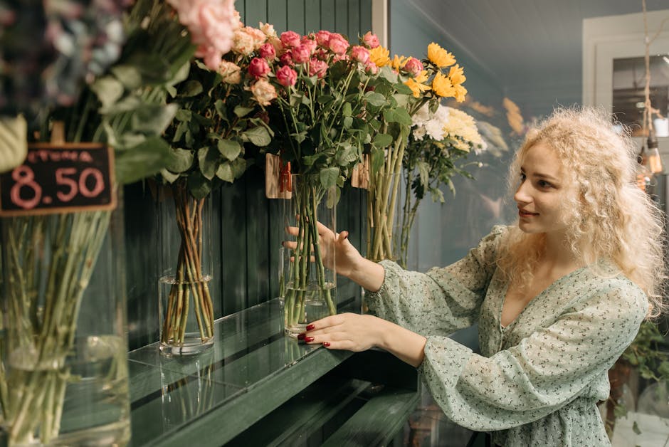 A woman with curly blonde hair and fair skin inspects a selection of fresh flowers arranged in tall glass vases on a dark green shelf at a florist shop, with vibrant pink, yellow, and white blooms inc